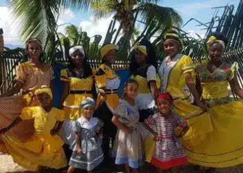 Personas vestidas con trajes tradicionales en una celebración cultural en la playa, rodeadas de palmeras y un cielo despejado. Foto vibrante que destaca la identidad cultural y la festividad en un entorno paradisíaco.