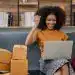 Entusiasta mujer migrante celebrando éxito con laptop y paquetes de envío en oficina moderna.