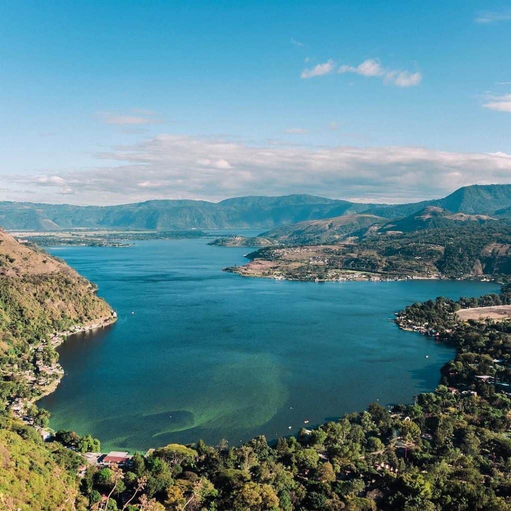 Paisaje de embalse en montaña, agua cristalina rodeada de vegetación y cerros con cielo azul, reflejando el entorno natural en una vista panorámica.