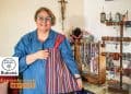 El arte de Silvia Patricia Flores hecho con historia alma y amor | Hecho en Guatemala – SoyMigrante.com REVISTA Smiling woman in blue denim shirt holds a striped handmade bag beside a jewelry display rack in a craft shop.