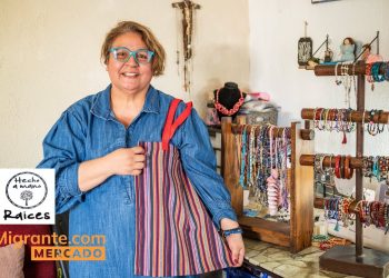 El arte de Silvia Patricia Flores hecho con historia alma y amor | Hecho en Guatemala – SoyMigrante.com REVISTA Smiling woman in blue denim shirt holds a striped handmade bag beside a jewelry display rack in a craft shop.