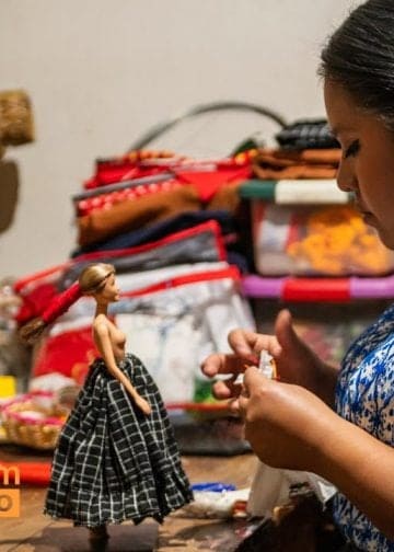 Muñecas que celebran la diversidad maya el sueño de Séfora – SoyMigrante.com REVISTA Guatemalan artisan sewing a doll in a busy fabric market, with 'SoyMigrante.com' logo in the corner