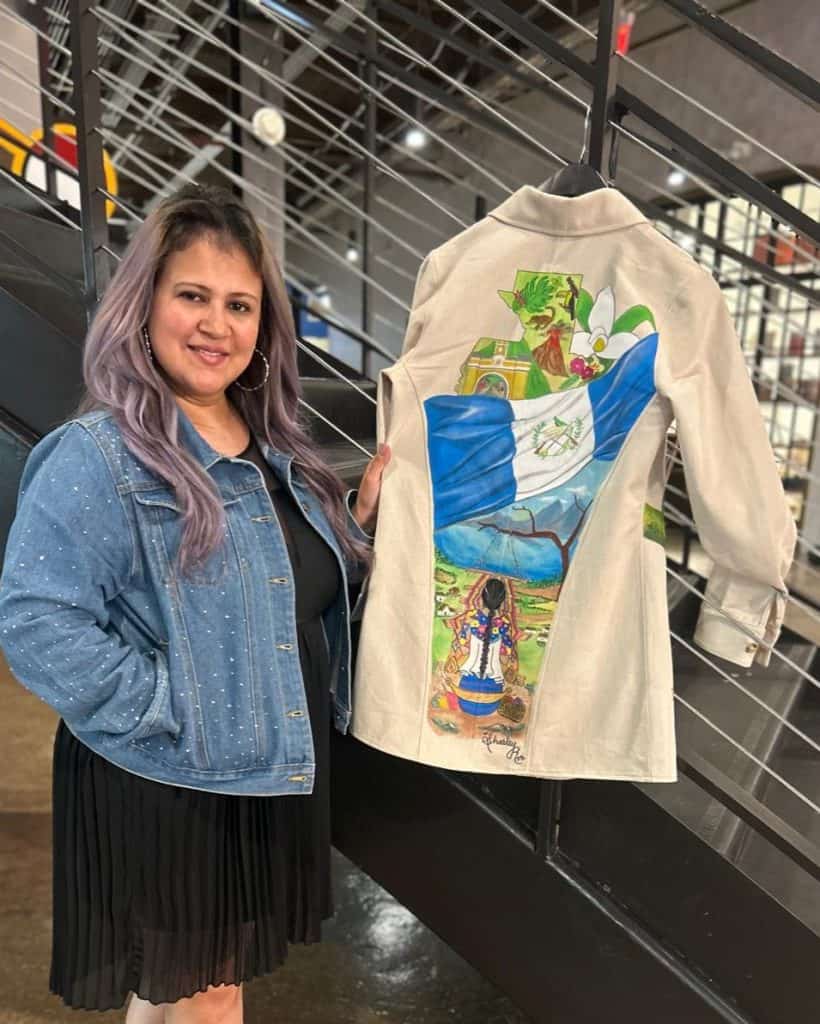 SoyMigrante.com REVISTA, obra de arte pintada a mano en chaqueta beige con paisaje, mujer indígena y símbolos culturales, resaltando identidad y migración, exhibida en evento cultural en ciudad moderna.