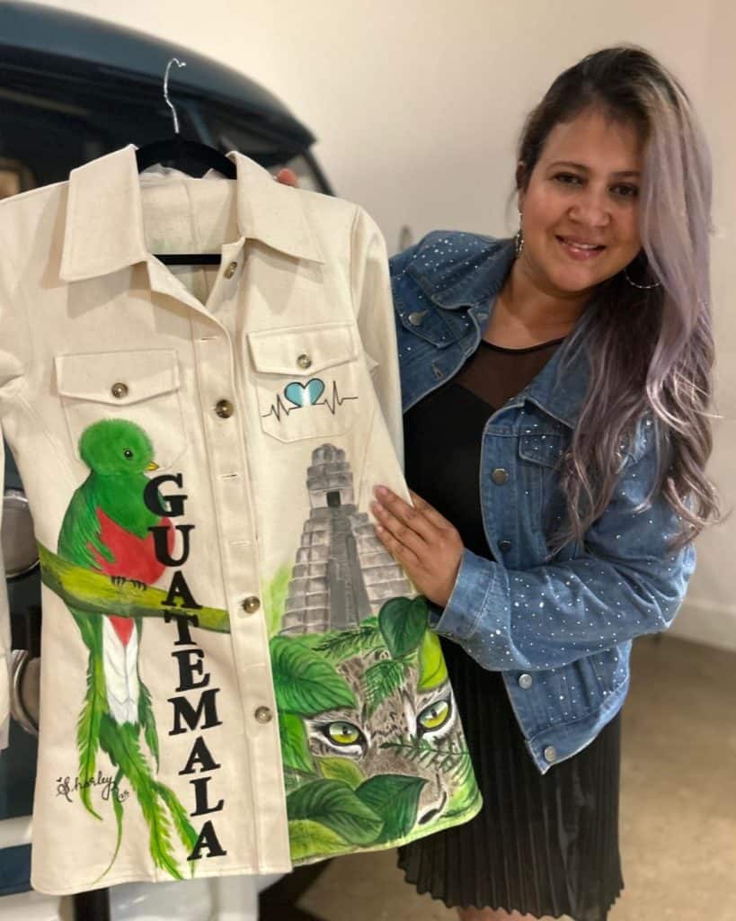Varias personas en un evento cultural, mostrando una chaqueta decorada con motivos guatemaltecos, arte y cultura, en el marco de la revista SoyMigrante.com REVISTA.