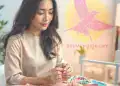 Jovenc woman with dark hair browsing colorful jewelry, cupcakes, and accessories on a table at a craft fair or market, in a bright, cheerful setting.