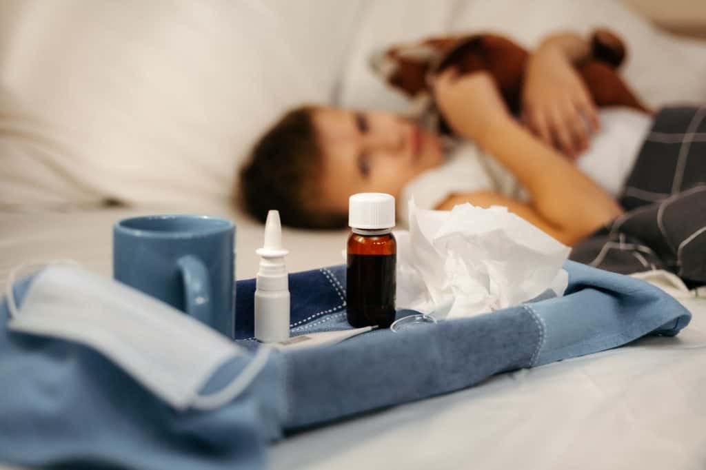 Fármaco y medicamentos sobre la cama, con una niña enferma recostada en el fondo, representando temas de salud, medicina y bienestar en contexto de migrantes. – SoyMigrante.com REVISTA