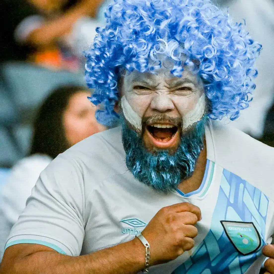 Disfrutando del fútbol con pasión en la Copa del Mundo, hincha con peluca y barba azules, expresando emoción y alegría en el estadio.