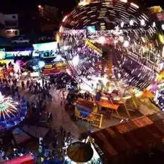 Espectáculo nocturno en feria con luces y atracciones, reflejo en una estructura metálica gigante que ilumina el evento. – SoyMigrante.com REVISTA