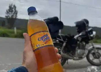 India Quiché la bebida que resiste al paso del tiempo en Guatemala – SoyMigrante.com REVISTA Refresco de crema soda en una mano, con motocicleta y paisaje en el fondo, representando la cultura y la experiencia del migrante en el recorrido.