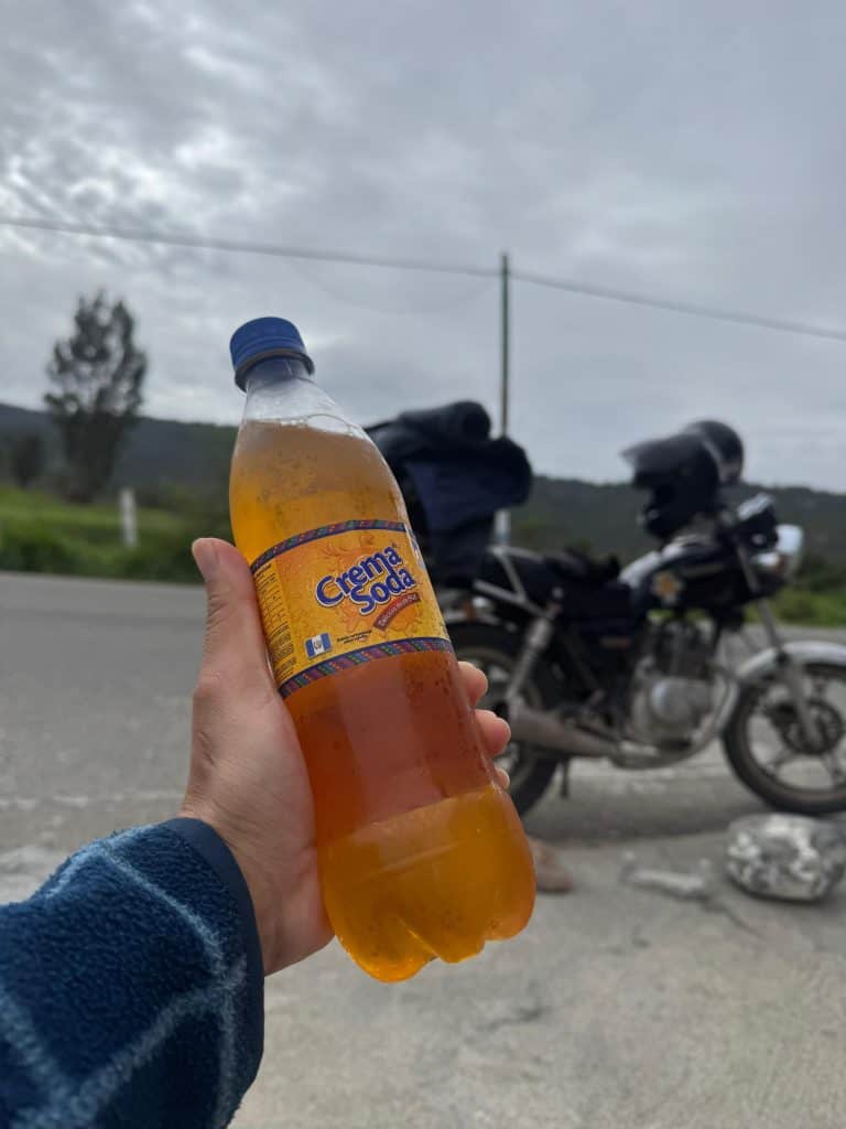 Refresco de crema soda en una mano, con motocicleta y paisaje en el fondo, representando la cultura y la experiencia del migrante en el recorrido.