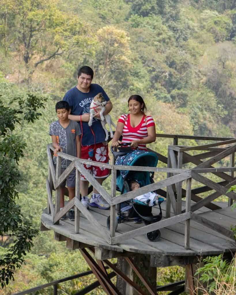 Caminatas seguras y al aire libre: los pulmones verdes de la ciudad de Guatemala – SoyMigrante.com REVISTA Personas disfrutando de un paseo por la naturaleza en un mirador de madera con vista a árboles y paisaje forestal. Familia con perros y niños en un entorno al aire libre, promoviendo el turismo y la vida saludable. – SoyMigrante.com REVISTA