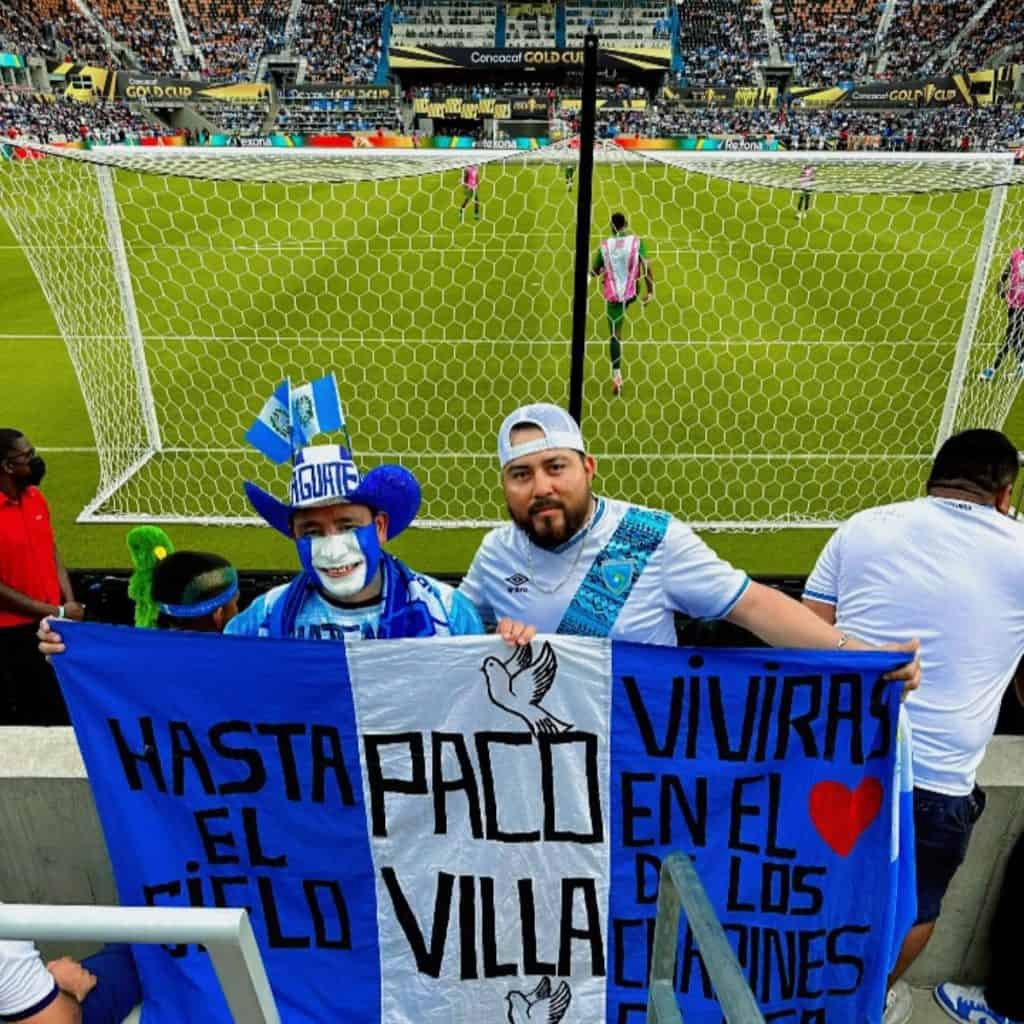 Jugadores de fútbol en estadio con aficionado con bandera de apoyo a El Salvador en partido internacional de fútbol en vivo - fotos de fútbol para revista soymigrante.com.