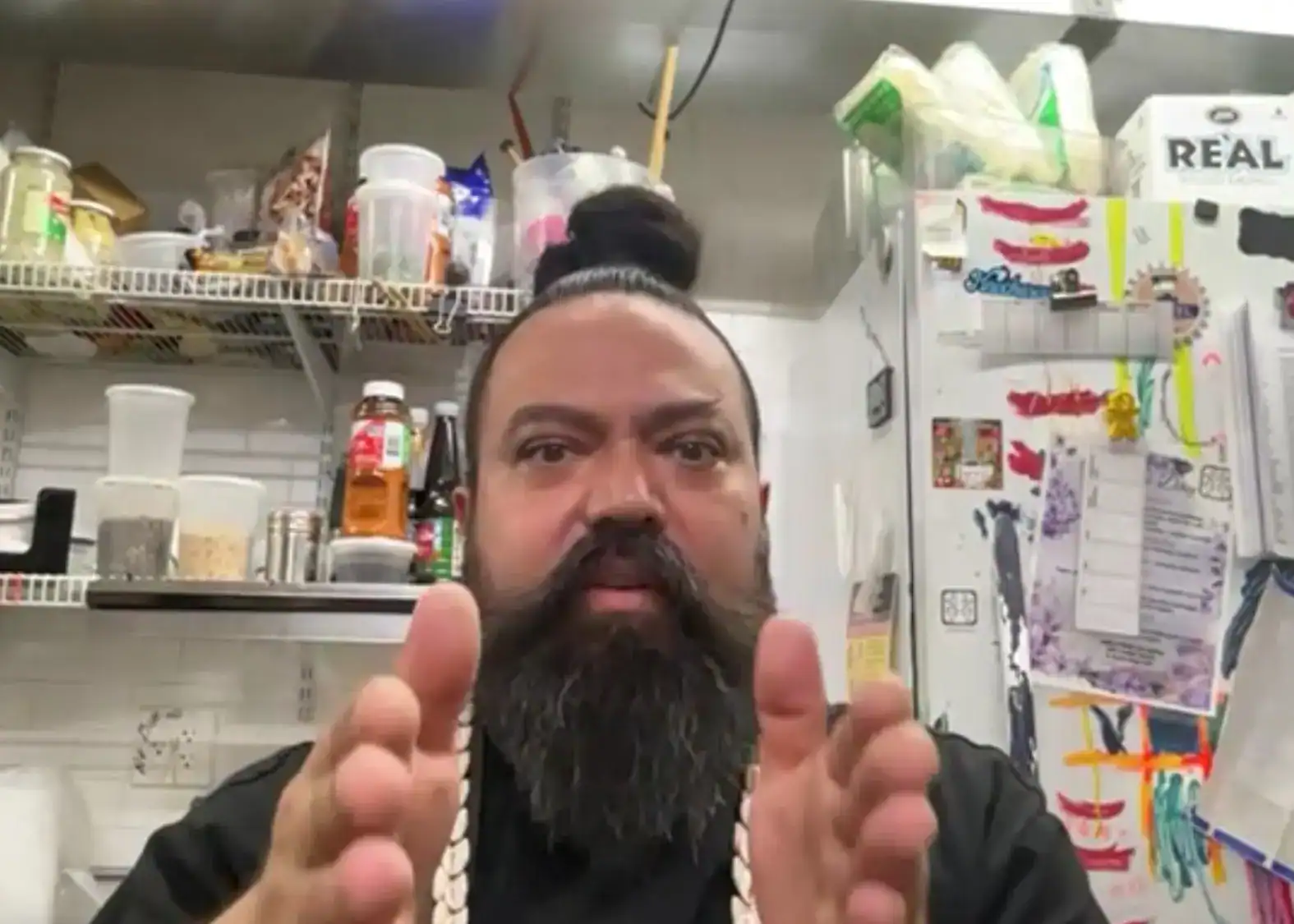 Barba, cabello peinado en coleta, fondo de despensa con alimentos y productos de cocina, hombre con cadena y gesto de enfado, estilo casual en contexto de comunidad migrante. – SoyMigrante.com REVISTA