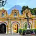Fachada de una iglesia colonial en Guatemala con montañas en el fondo, resaltando su arquitectura colorida y historia cultural.
