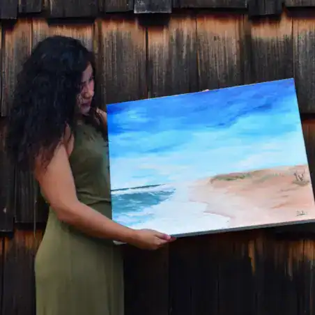 Cargando una pintura de playa al óleo en un jardín, destacando arte y cultura migrante en SoyMigrante.com REVISTA para promover comunidades creativas y diversidad cultural.