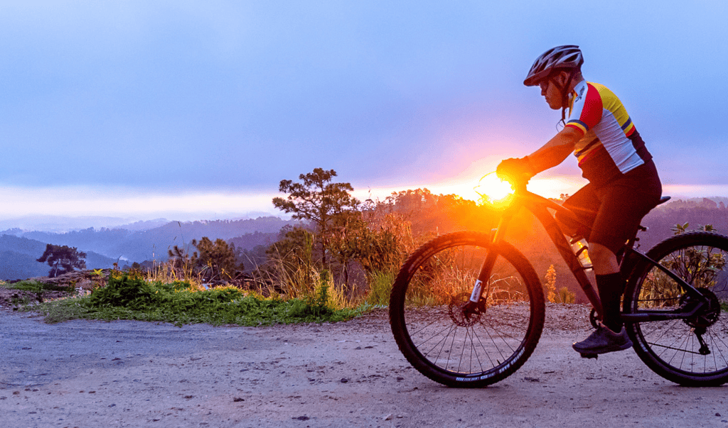 Andar en bicicleta al atardecer, deportista en ruta rural, actividad al aire libre para promoción de vida saludable y turismo sostenible, con paisaje de montañas y naturaleza. – SoyMigrante.com REVISTA