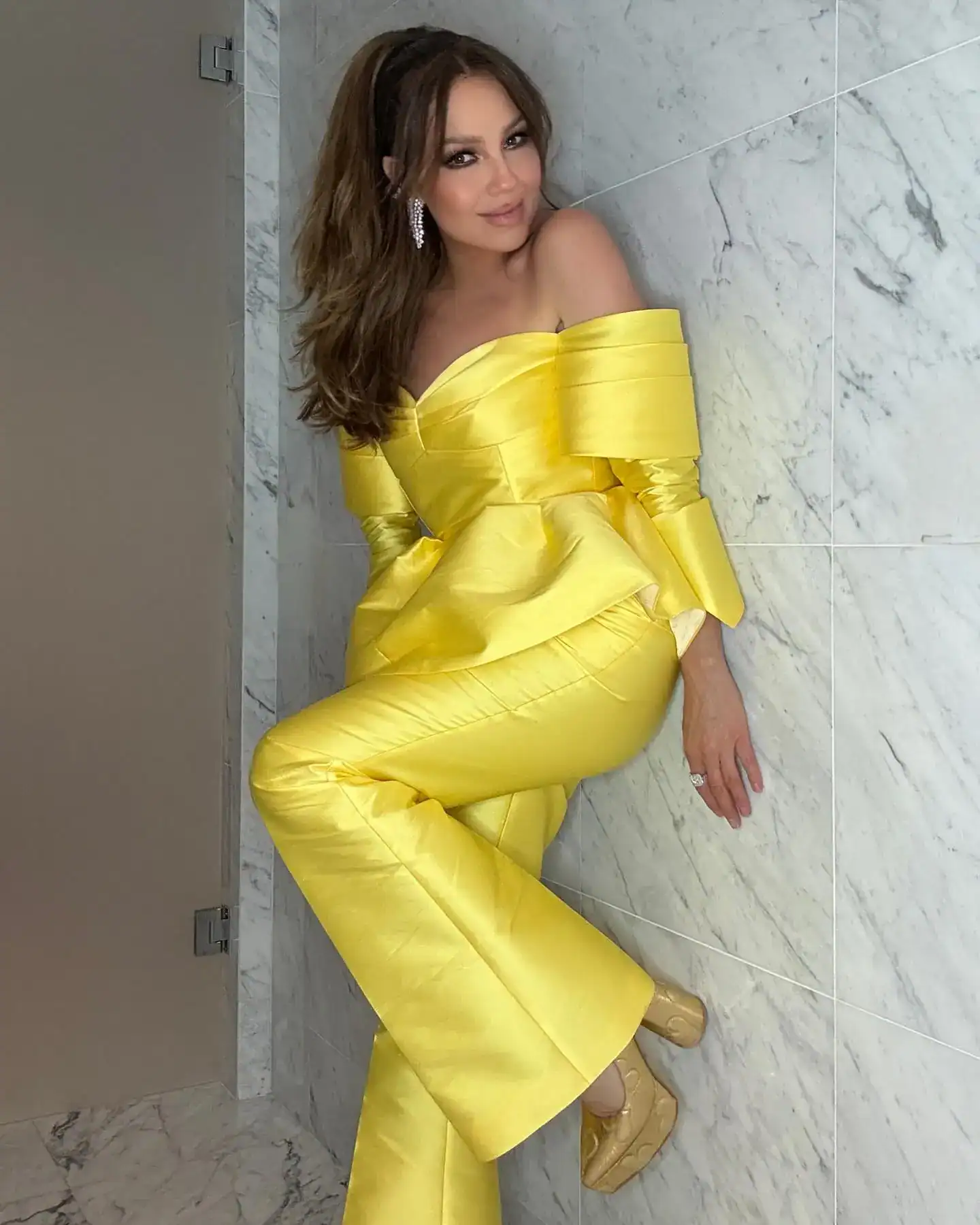 Artistas y figuras públicas alzan la voz en apoyo a migrantes ante redadas y trato injusto – SoyMigrante.com REVISTA Vestido amarillo brillante de gala, mujer posando en un fondo de paredes de mármol, estilo elegante y sofisticado, revista SoyMigrante.com, enfoque en moda y cultura migrante en español.