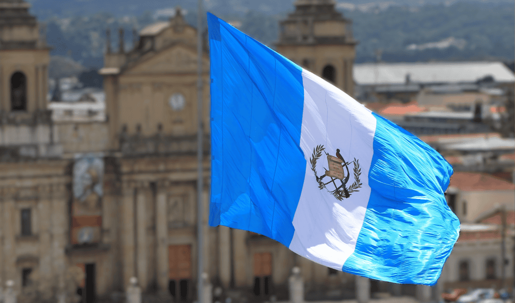 Bandera de Guatemala ondeando en ciudad histórica, símbolo de identidad y cultura en Latinoamérica para artículo sobre migrantes y comunidad guatemalteca. – SoyMigrante.com REVISTA