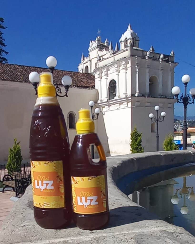 Dos botellas de miel LUZ en primer plano, sobre un muro de piedra en un entorno urbano con iglesia colonial blanca y farolas antiguas, simbolizando tradición y cultura en México. – SoyMigrante.com REVISTA