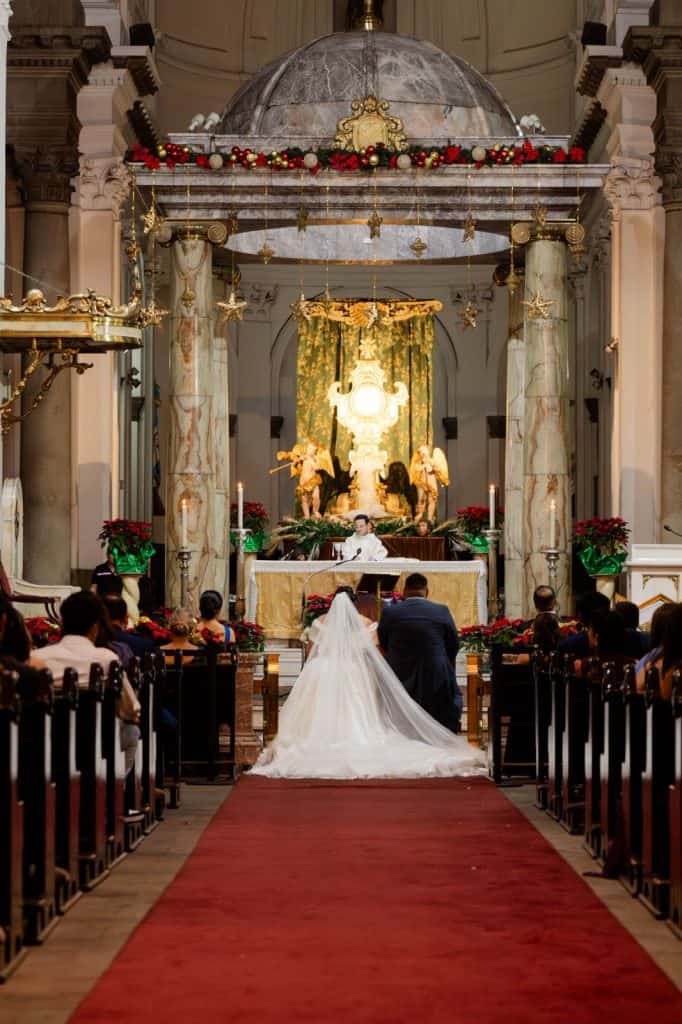 Boda religiosa en una iglesia elegante con decoración navideña, altar adornado con velas y flores rojas, ceremonia matrimonial tradicional con novios y asistentes. – SoyMigrante.com REVISTA