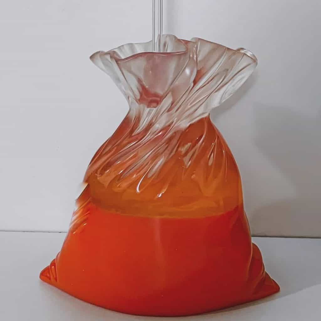 Bote de vidrio con líquido naranja, envuelto en plástico transparente, sobre fondo blanco. – SoyMigrante.com REVISTA
