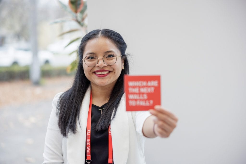 Participante latina con gafas sonriendo y sosteniendo una tarjeta roja con mensaje inspirador, en un evento de la comunidad migrante, promoviendo la inclusión y el empoderamiento en torno a temas de migración y participación social. – SoyMigrante.com REVISTA
