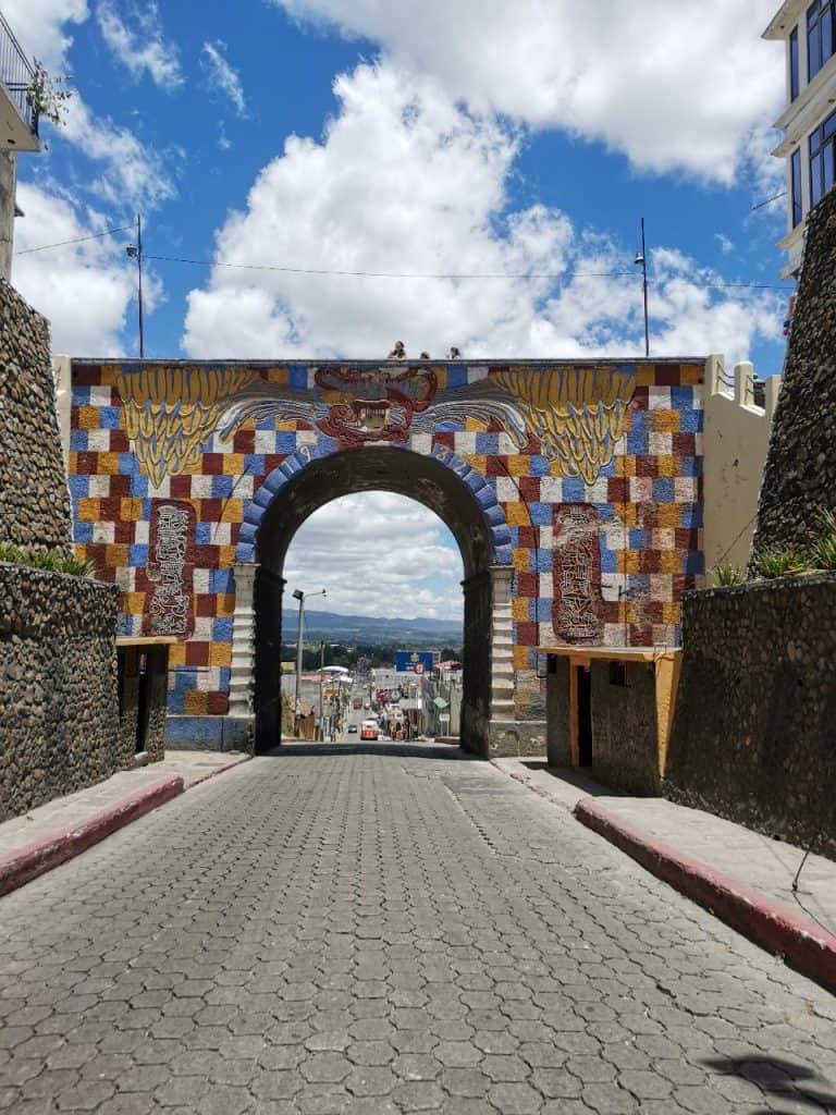 Puerta monumental en un callejón empedrado con mural colorido en la entrada, cielo despejado, vista de la ciudad y paisaje en el fondo, relevante para artículos sobre cultura y arquitectura mexicana. – SoyMigrante.com REVISTA