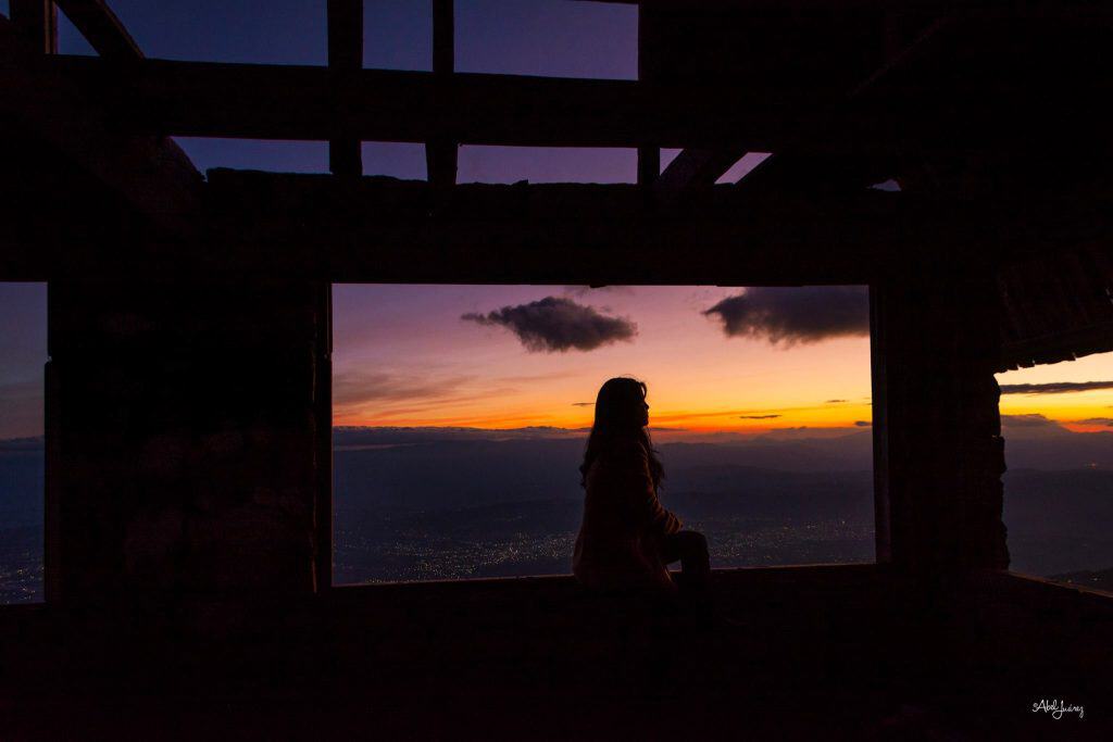 Atardecer sobre las montañas con mujer sentada contemplando el paisaje, en una estructura de madera, resaltando la cultura migrante en México en un entorno natural.
