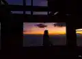 Atardecer sobre las montañas con mujer sentada contemplando el paisaje, en una estructura de madera, resaltando la cultura migrante en México en un entorno natural. – SoyMigrante.com REVISTA