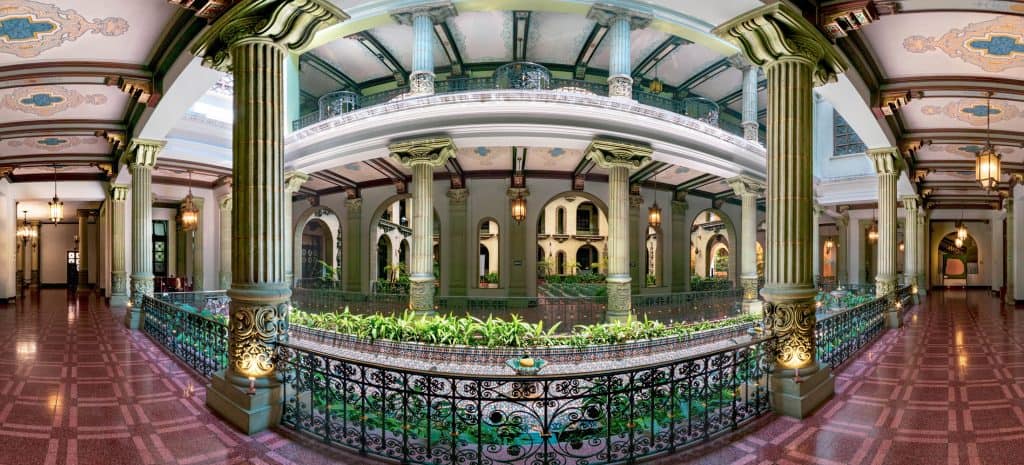 Imágenes del interior de un edificio histórico con columnas clásicas, decoraciones detalladas y un exuberante jardín central, reflejando arquitectura elegante y cultural para la comunidad migrante. – SoyMigrante.com REVISTA