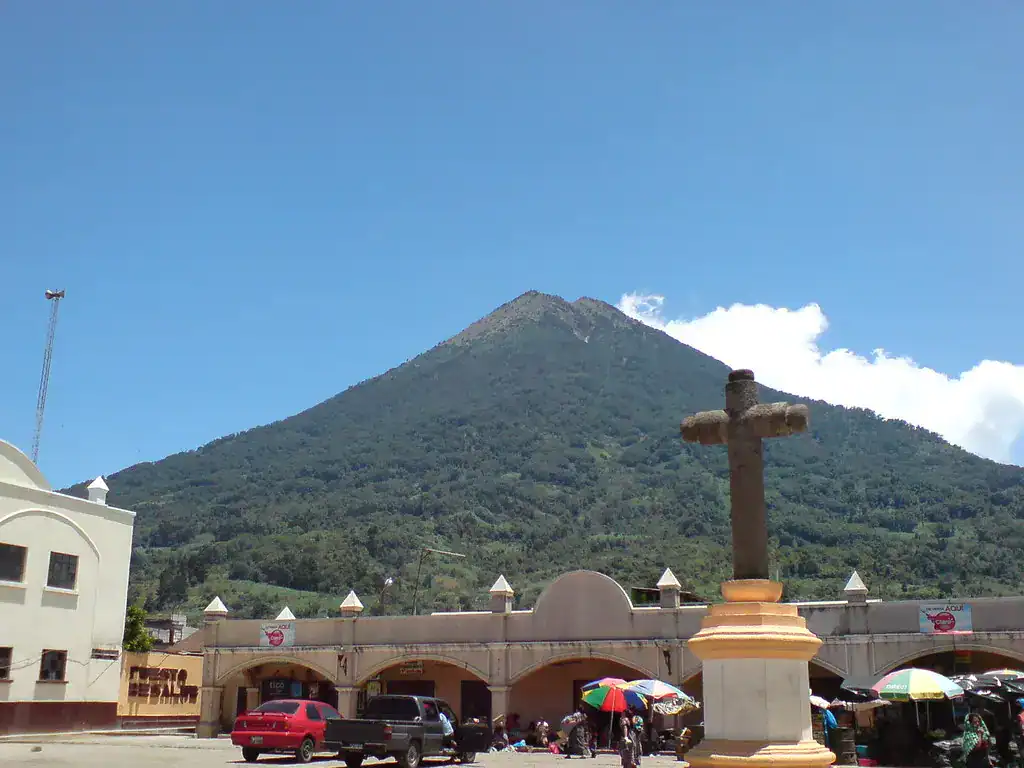 Montaña volcánica en el fondo, iglesia y plaza en primer plano, con puestos de mercado y personas bajo paraguas coloridos en un día soleado en un pueblo mexicano. – SoyMigrante.com REVISTA