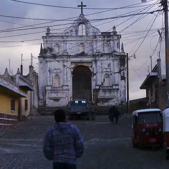 Camino hacia una iglesia colonial en una calle empedrada, con personas caminando y un auto en el fondo, en un día nublado. – SoyMigrante.com REVISTA