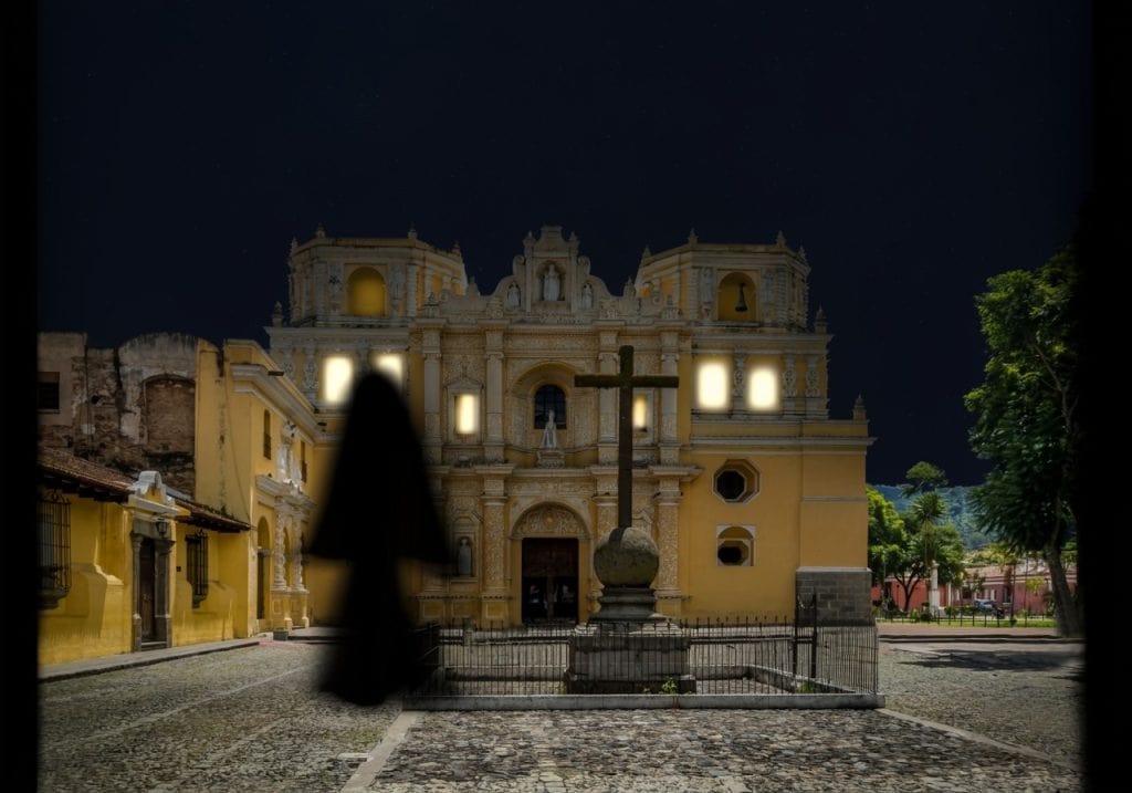 Catedral de noche en un pueblo colonial con estructura histórica en el centro, iluminada, con una cruz en primer plano y árboles a su alrededor. – SoyMigrante.com REVISTA