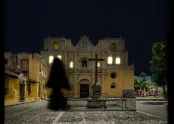 Catedral de noche en un pueblo colonial con estructura histórica en el centro, iluminada, con una cruz en primer plano y árboles a su alrededor.