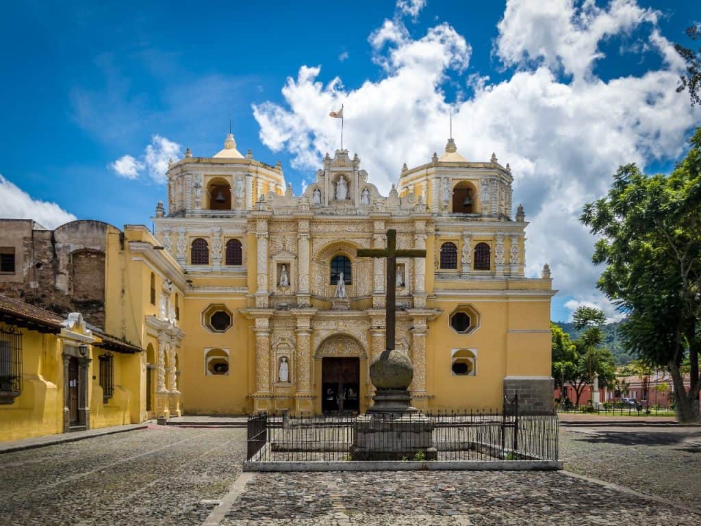 Arco Iglesia colonial en colores amarillos y detalles ornamentales en un cielo nublado, rodeada de árboles y calles empedradas, reflejando la arquitectura histórica de Guatemala. – SoyMigrante.com REVISTA