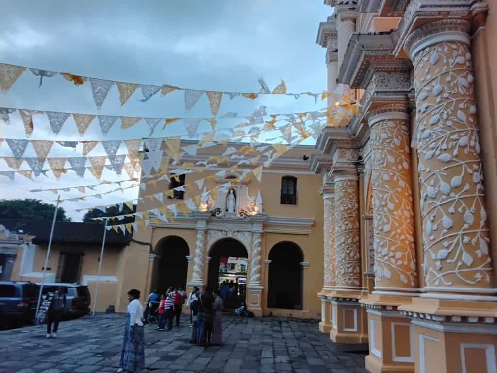 Fachada de iglesia colonial decorada con banderines blancos y amarillos en una plaza, con personas reunidas, cielo nublado, arquitectura historicista, fotografía urbana en México, enfoque en detalles ornamentales y cultura local. – SoyMigrante.com REVISTA