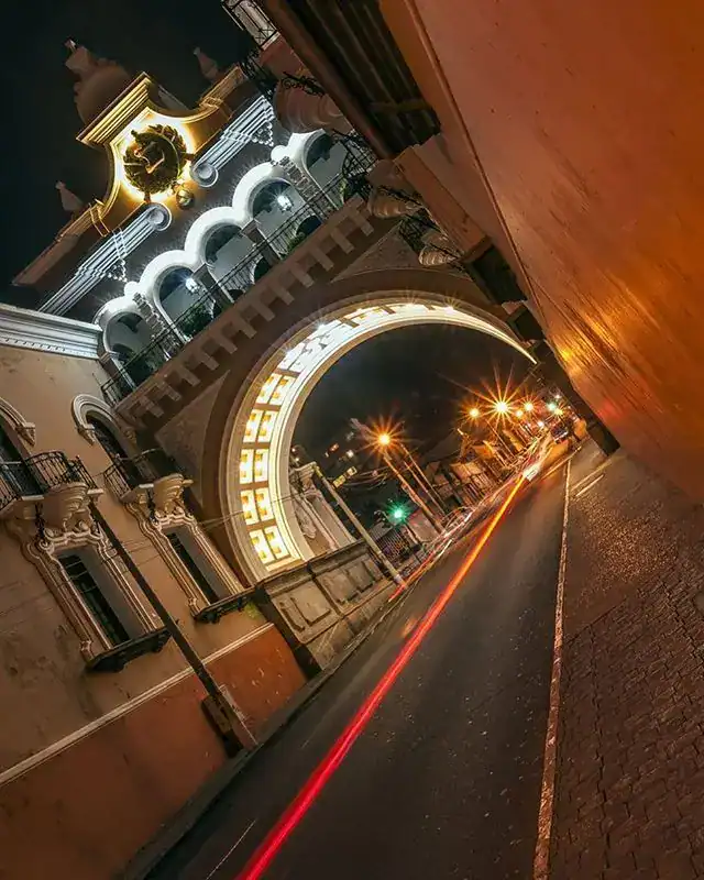 Puerta de la Ciudad de México iluminada de noche con luces decorativas y tráfico en movimiento. – SoyMigrante.com REVISTA