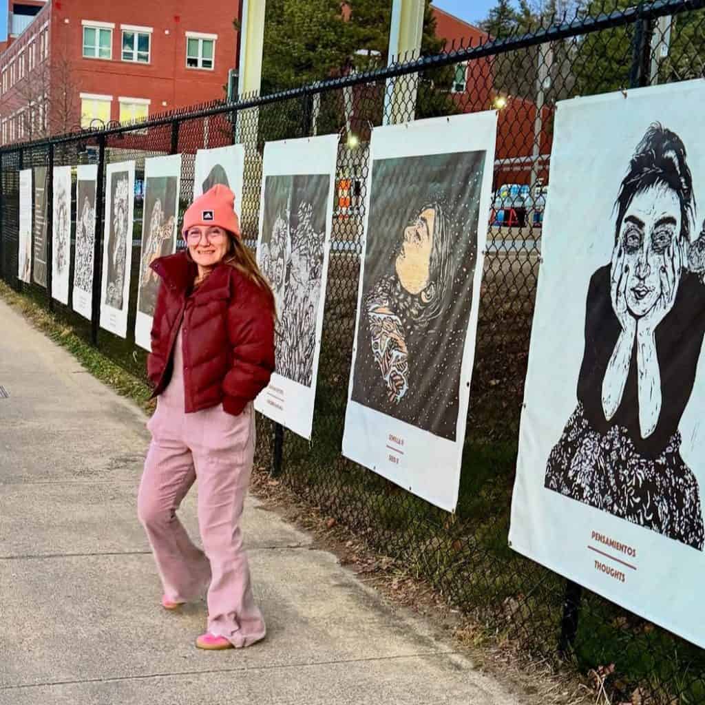 Fotografía de una mujer sonriente en una exposición al aire libre con retratos en blanco y negro en una valla de malla, destacando arte, cultura y creatividad en el contexto de SoyMigrante.com REVISTA.