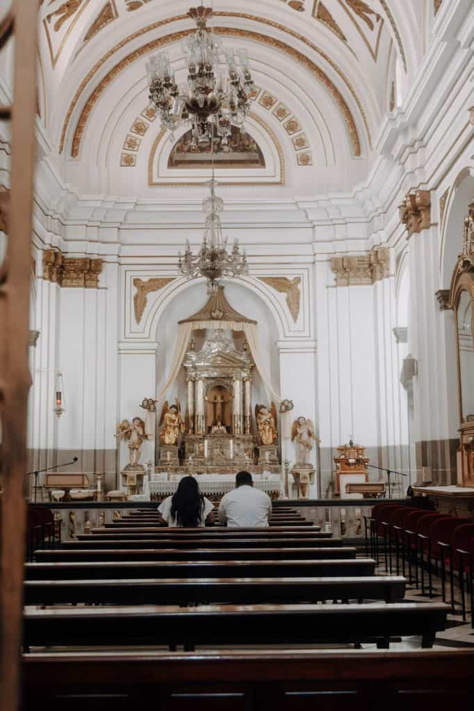 Alto texto: Interior de una iglesia con arquitectura clásica, columnas blancas, altar ornamentado y candelabros elegantes, luminosa y decorada en estilo barroco. – SoyMigrante.com REVISTA