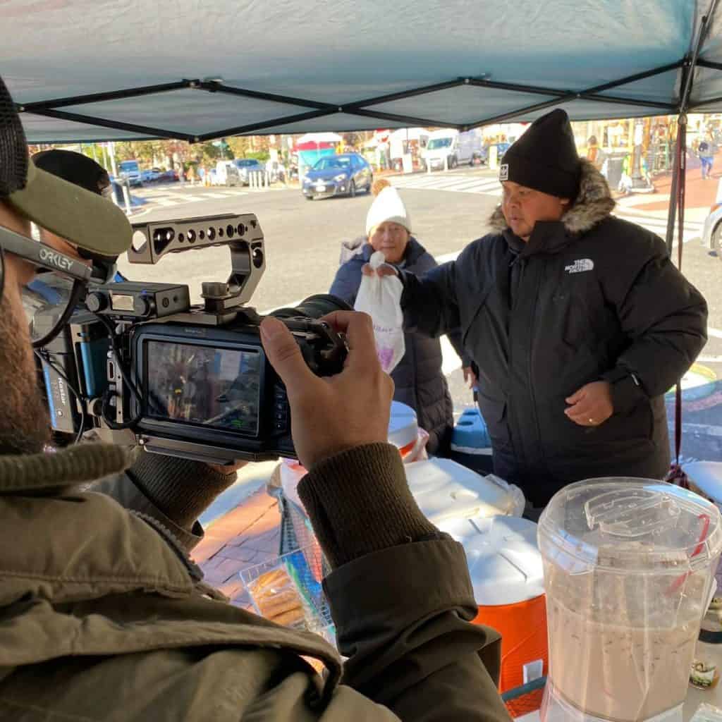 Cámara profesional grabando a un vendedor en mercado callejero durante una entrevista o reportaje, promoviendo el contenido visual de SoyMigrante.com REVISTA sobre migrantes y comunidades. – SoyMigrante.com REVISTA