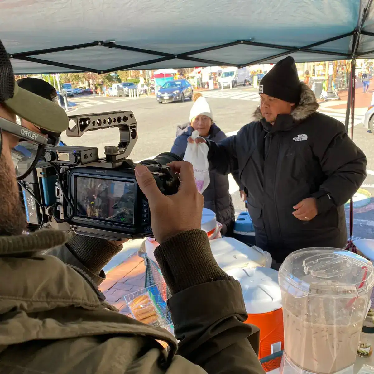 cocinando guatemala serie restaurantes chapines en USA 2 – SoyMigrante.com REVISTA Cámara profesional grabando a un vendedor en mercado callejero durante una entrevista o reportaje, promoviendo el contenido visual de SoyMigrante.com REVISTA sobre migrantes y comunidades.
