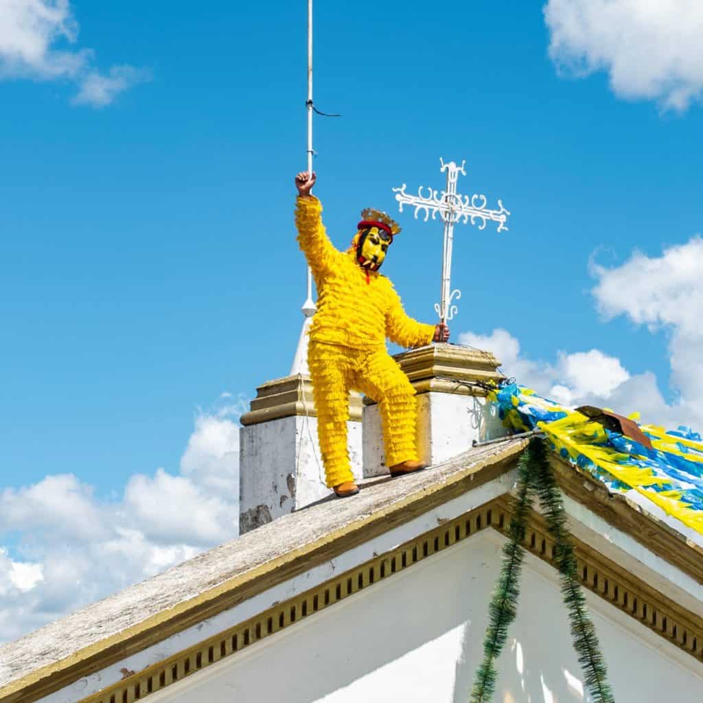 Figura decorativa en el techo de una iglesia en una celebración tradicional, vestida con disfraz amarillo y máscara, sosteniendo un símbolo religioso, en un día soleado con cielo azul y nubes. – SoyMigrante.com REVISTA