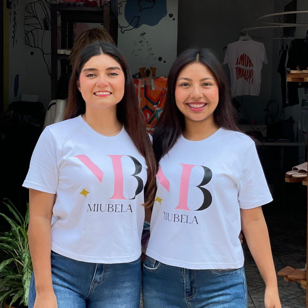 Chicas sonriendo en promoción de Mueblea con camisetas blancas con logotipo en un negocio de muebles, mostrando diversidad y emprendimiento femenino. – SoyMigrante.com REVISTA
