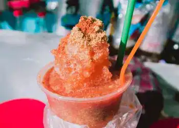 Refresco de hielo picado con sabor en vaso transparente, envuelto en bolsa plástica, con popotes verdes y naranjas, en un puesto callejero, típico de la gastronomía mexicana y de migrantes.