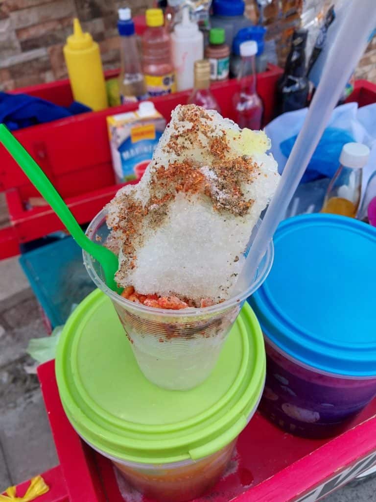 Refresco de hielo raspado con sabor a frutas y toppings en vaso transparente, en un puesto callejero, rodeado de bottles de condimentos y utensilios para comida. – SoyMigrante.com REVISTA