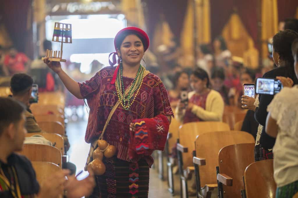 Artesana indígena durante evento cultural con vestimenta tradicional en un auditorio lleno de asistentes tomando fotos.