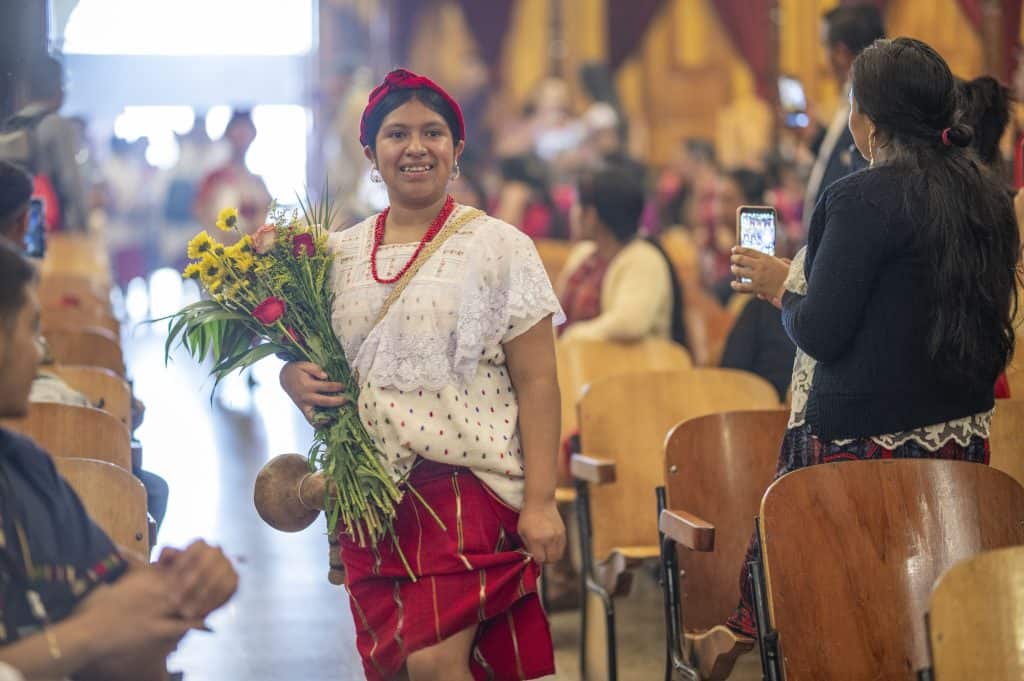 Niña con traje tradicional y flores en mano en evento cultural indígena en iglesia.