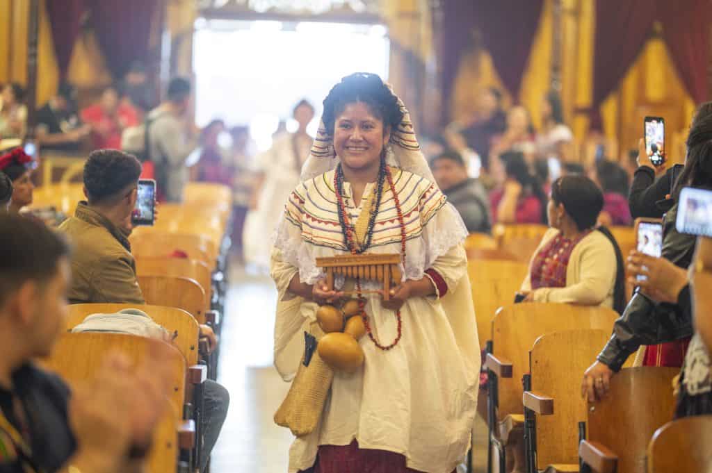 Tradicional mujer indígena con vestimenta cultural en evento en un espacio cerrado lleno de asistentes tomando fotos.