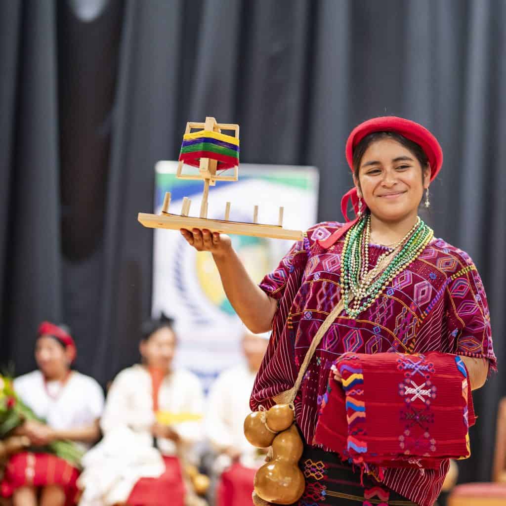 Colombiana vestida con traje tradicional indígena, sosteniendo una figura en miniatura de una embarcación artesanal, participando en evento cultural con fondo de cortinas negras, promoviendo cultura y tradiciones ancestrales.