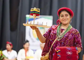 Colombiana vestida con traje tradicional indígena, sosteniendo una figura en miniatura de una embarcación artesanal, participando en evento cultural con fondo de cortinas negras, promoviendo cultura y tradiciones ancestrales.